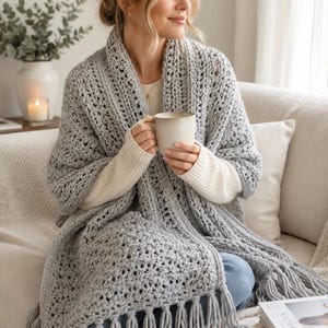 May include: A light grey crochet shawl featuring a textured stitch pattern and long fringe along the bottom edge. The shawl is draped over the shoulders and upper body, showcasing the intricate yarn work and the soft, open weave of the fabric.