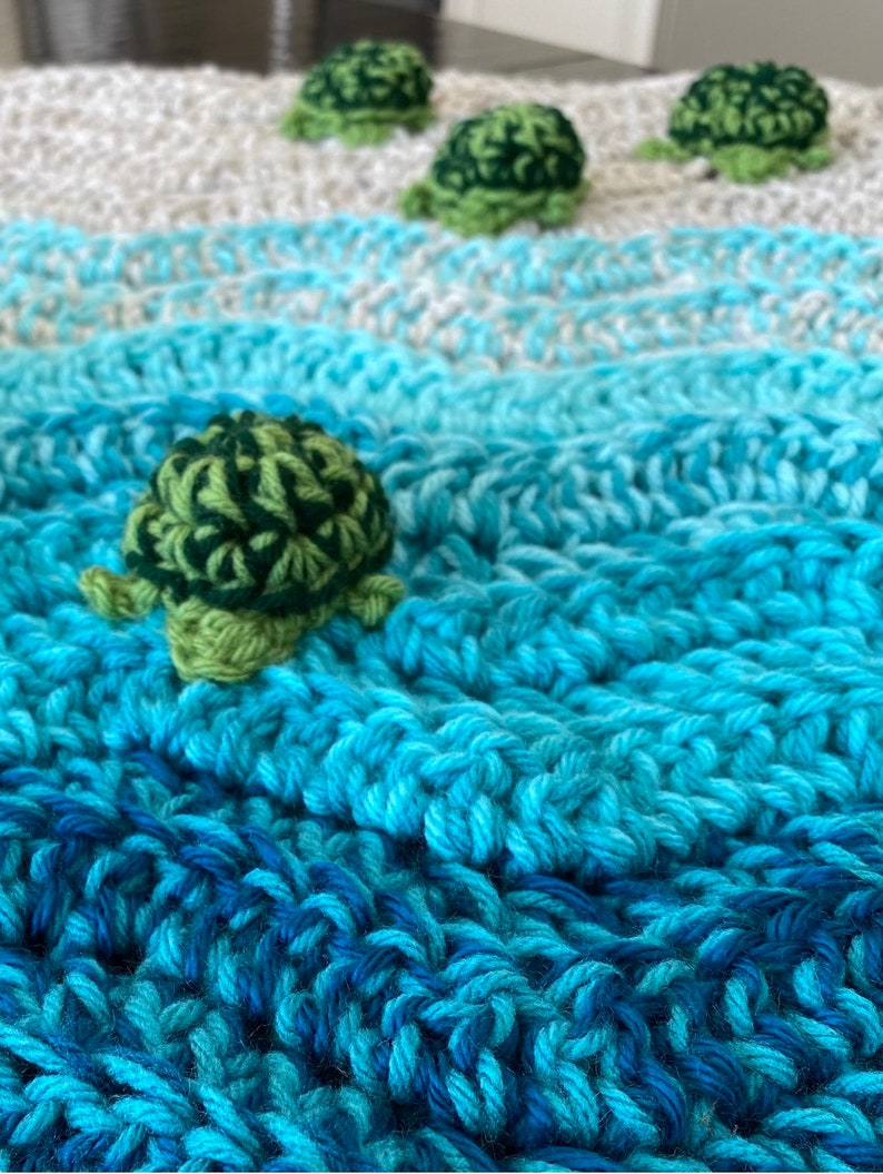 May include: A handmade crochet blanket in shades of blue, white, and gray, resembling ocean waves. Several small, green and dark green crochet turtles are scattered across the blanket, adding a playful touch. The blanket is made with thick yarn.