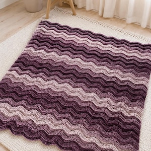 May include: A rectangular crochet blanket featuring a wavy ripple pattern in a gradient of purple shades, transitioning from deep dark plum to lighter lavender and soft mauve tones throughout the textured, hand-stitched fabric construction.