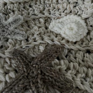 May include: Close-up of a handmade crochet blanket with a textured surface. The blanket features a mix of cream, light blue, and brown yarn. Decorative elements include star and leaf shapes, adding a unique touch to the cozy textile.