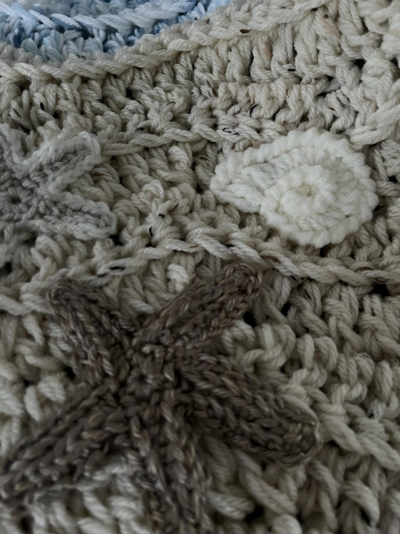 May include: Close-up of a handmade crochet blanket with a textured surface. The blanket features a mix of cream, light blue, and brown yarn. Decorative elements include star and leaf shapes, adding a unique touch to the cozy textile.