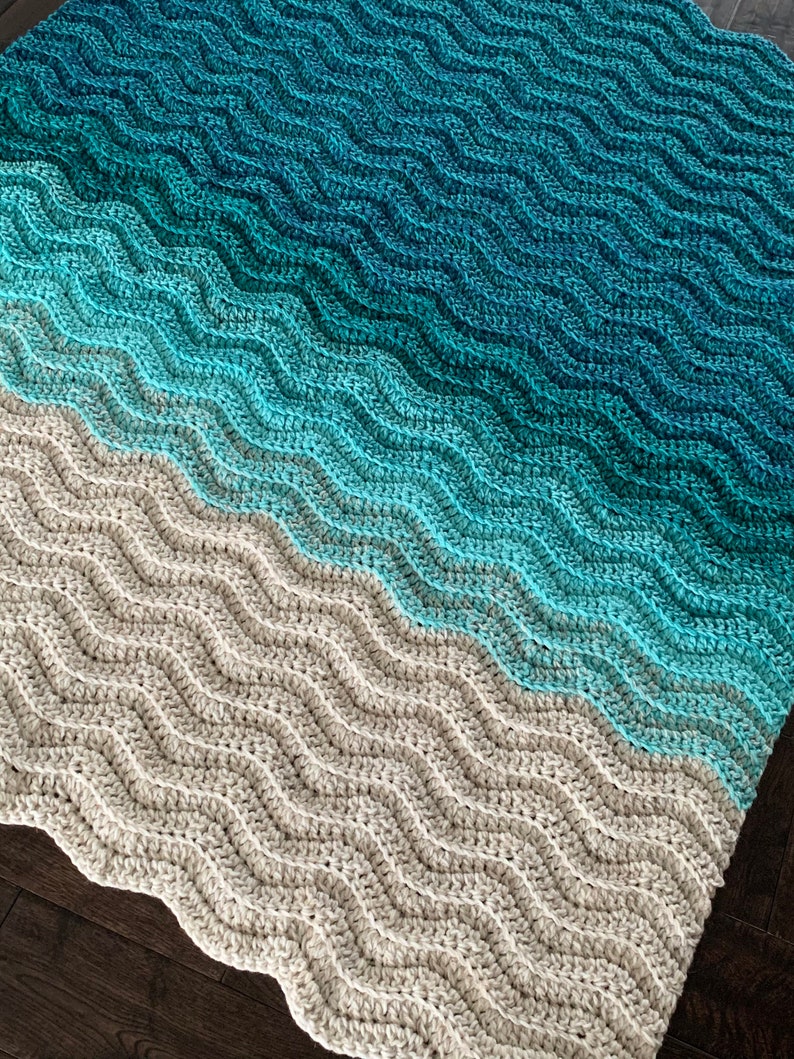 May include: A handmade crochet blanket with a wave pattern. The blanket transitions from a light gray at the bottom to shades of blue and teal at the top. The wavy design adds texture and visual interest.