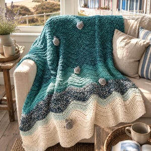 May include: A handmade crochet blanket in shades of teal, blue, and cream, resembling ocean waves. The blanket is draped over a beige sofa and adorned with gray pom-poms. The scene includes a coastal view through a window.
