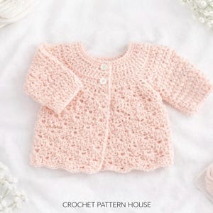 May include: A light pink, crocheted baby cardigan with long sleeves and two white buttons. The scalloped hem and textured stitch create a delicate look. The text "CROCHET PATTERN HOUSE" is at the bottom.