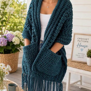 May include: A dark teal crochet cardigan featuring a chunky textured stitch, an open front design, two large patch pockets, and long fringe detailing along the bottom hem. The garment is worn over a white top and blue denim jeans.