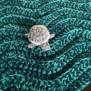 May include: A handmade crochet blanket in shades of teal and turquoise, featuring a textured chevron pattern. A small, gray and white crocheted turtle is placed on top of the blanket. The blanket is a cozy, handmade item.