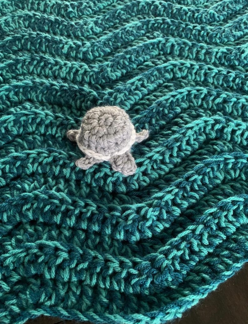 May include: A handmade crochet blanket in shades of teal and turquoise, featuring a textured chevron pattern. A small, gray and white crocheted turtle is placed on top of the blanket. The blanket is a cozy, handmade item.
