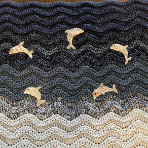 May include: A handmade crochet blanket with a wavy pattern in shades of blue, gray, and white, resembling ocean waves. Five beige dolphin appliqu&eacute;s are scattered across the blanket, adding a playful touch to the nautical theme.