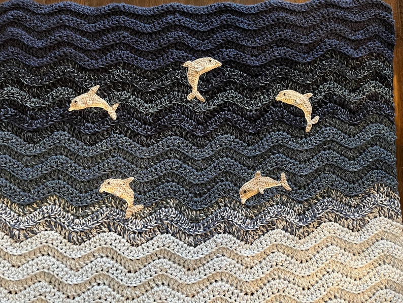 May include: A handmade crochet blanket with a wavy pattern in shades of blue, gray, and white, resembling ocean waves. Five beige dolphin appliqu&eacute;s are scattered across the blanket, adding a playful touch to the nautical theme.