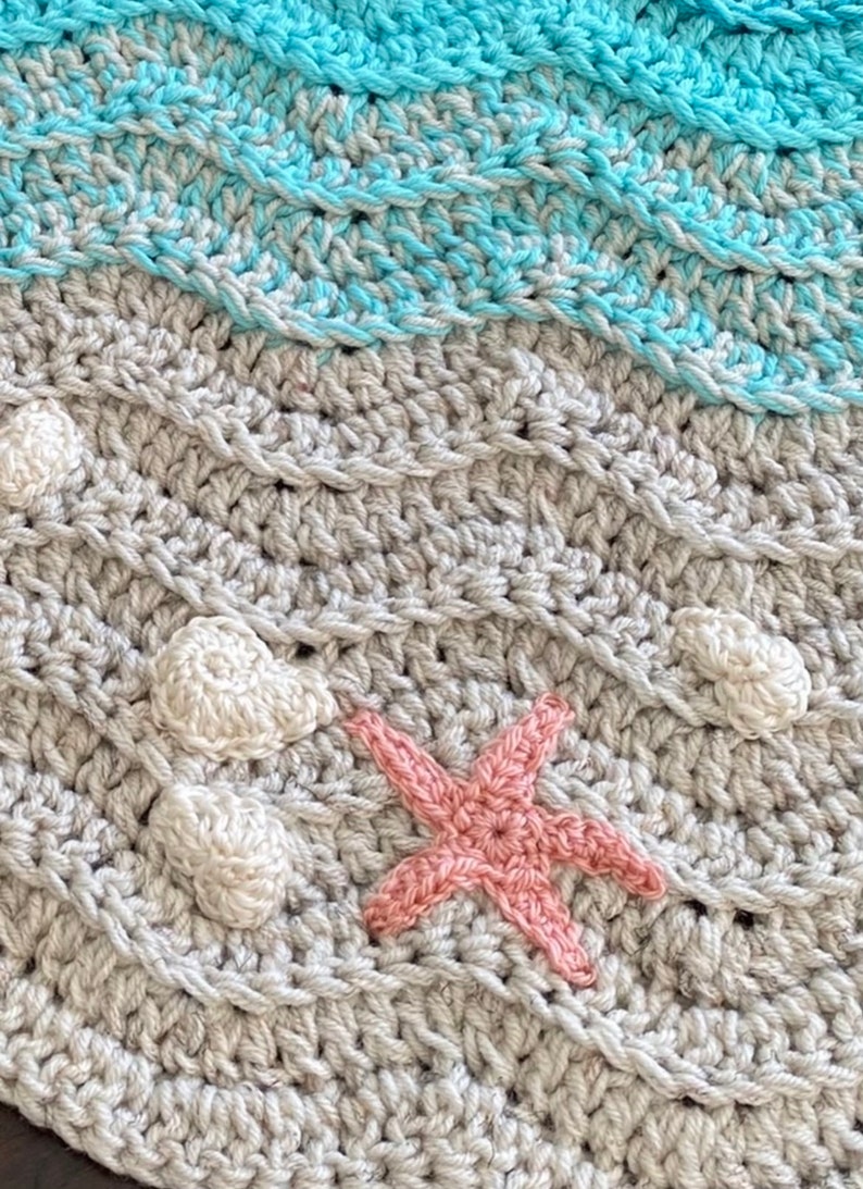 May include: Handmade crochet blanket with a beach theme. The blanket features a textured design with waves in shades of blue and gray, along with white seashell accents and a pink starfish. The blanket is a cozy and decorative item.