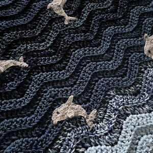 May include: A crocheted blanket with a wavy pattern in shades of blue, resembling ocean waves. Several gray crocheted dolphins are scattered across the blanket. The blanket is made of soft yarn and has a textured appearance.