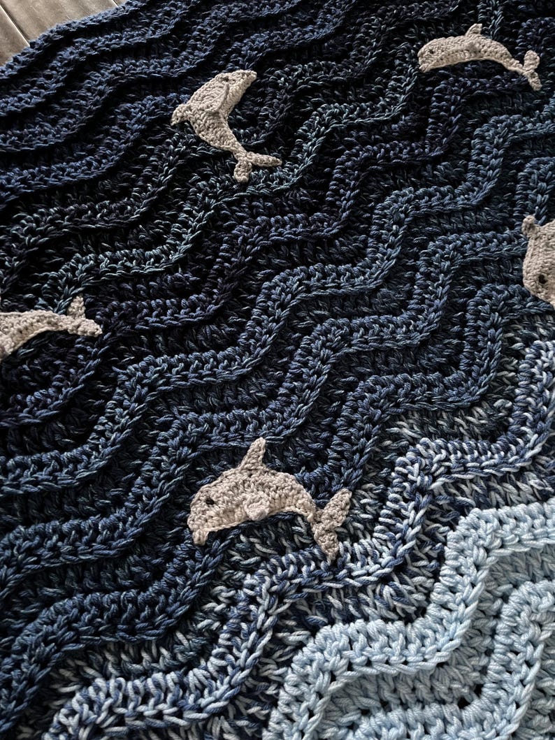 May include: A crocheted blanket with a wavy pattern in shades of blue, resembling ocean waves. Several gray crocheted dolphins are scattered across the blanket. The blanket is made of soft yarn and has a textured appearance.