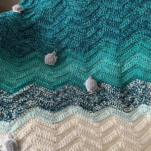 May include: A handmade crochet blanket with a sea-themed design. The blanket features a wave pattern in shades of teal, blue, and white, with small gray turtle appliqu&eacute;s scattered across the surface. The blanket is made of soft yarn.