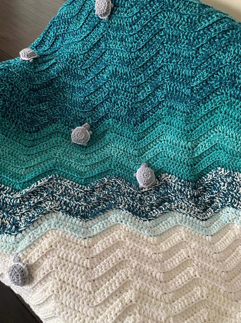 May include: A handmade crochet blanket with a sea-themed design. The blanket features a wave pattern in shades of teal, blue, and white, with small gray turtle appliqu&eacute;s scattered across the surface. The blanket is made of soft yarn.