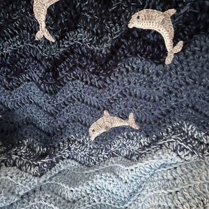 May include: A crocheted blanket with a wavy pattern in shades of blue, resembling ocean waves. Three gray crocheted dolphins are appliqued on the blanket, creating a nautical theme. The blanket is handmade and features a textured surface.