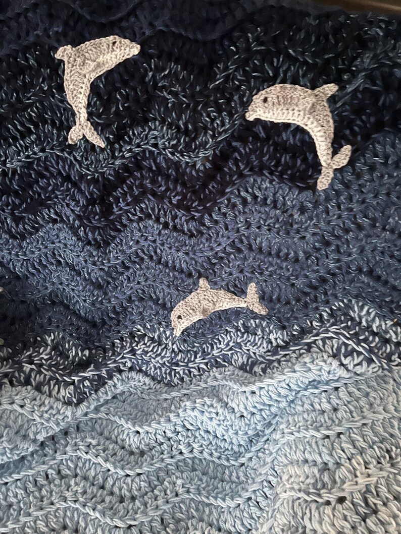May include: A crocheted blanket with a wavy pattern in shades of blue, resembling ocean waves. Three gray crocheted dolphins are appliqued on the blanket, creating a nautical theme. The blanket is handmade and features a textured surface.