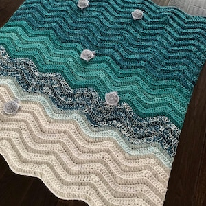 May include: A handmade crochet blanket with a beach-themed design. The blanket features a wave pattern in shades of teal, blue, and white, with small gray turtle accents. The blanket is made of soft yarn and is perfect for a cozy home decor.