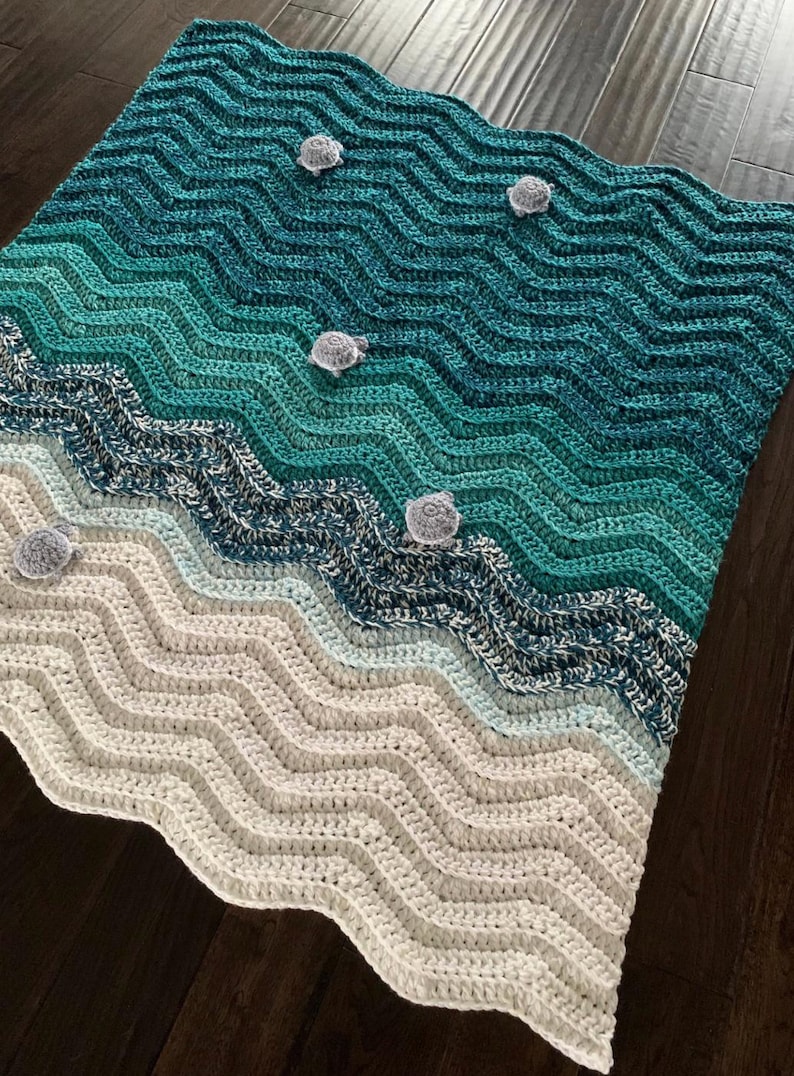 May include: A handmade crochet blanket with a beach-themed design. The blanket features a wave pattern in shades of teal, blue, and white, with small gray turtle accents. The blanket is made of soft yarn and is perfect for a cozy home decor.