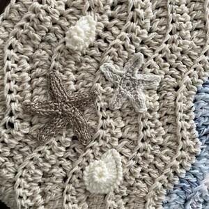 May include: Crocheted blanket in cream, tan, and blue hues, adorned with three-dimensional starfish and shell embellishments. The blanket features a textured stitch pattern, creating a cozy and decorative item. The color palette evokes a beach theme.