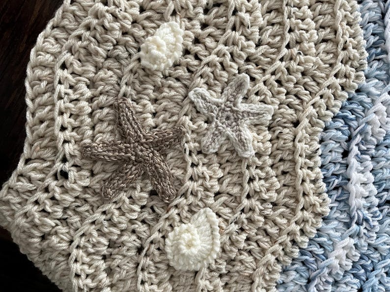 May include: Crocheted blanket in cream, tan, and blue hues, adorned with three-dimensional starfish and shell embellishments. The blanket features a textured stitch pattern, creating a cozy and decorative item. The color palette evokes a beach theme.