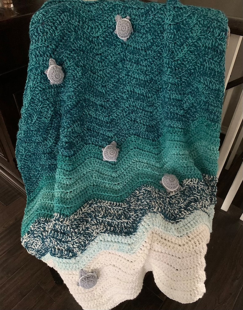 May include: A handmade crochet blanket in shades of teal, green, and white, resembling ocean waves. The blanket features several gray turtle appliqu&eacute;s. The blanket is draped over a dark surface.