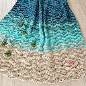 May include: A handmade crochet blanket depicting a beach scene. The blanket features wavy patterns in shades of blue, turquoise, and beige, with green pom-poms, white shells, and a pink starfish. The blanket is made of soft yarn.