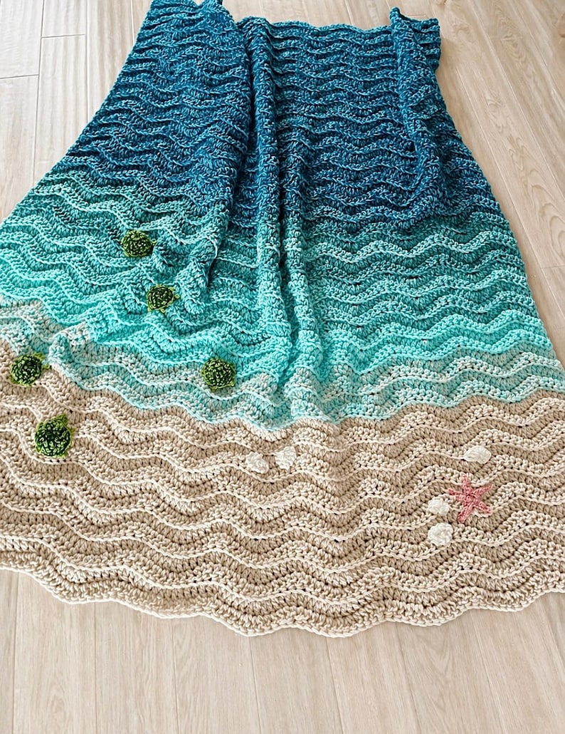 May include: A handmade crochet blanket depicting a beach scene. The blanket features wavy patterns in shades of blue, turquoise, and beige, with green pom-poms, white shells, and a pink starfish. The blanket is made of soft yarn.