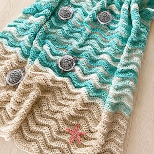 May include: A handmade crochet blanket with a beach theme. The blanket features a wavy pattern in shades of blue, beige, and white, with appliqued sea turtles and a pink starfish. The blanket is approximately 60 inches long.