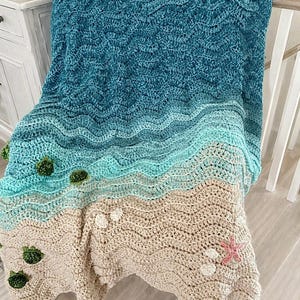 May include: A handmade crochet blanket with a beach theme. The blanket features a gradient of blues and greens representing the ocean, transitioning to a tan color for the sand. Embellishments include green seaweed and white seashells, with a pink starfish.
