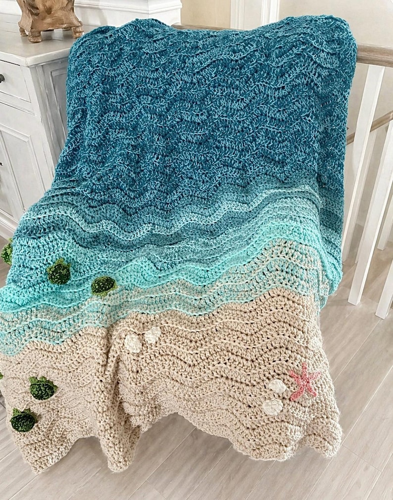 May include: A handmade crochet blanket with a beach theme. The blanket features a gradient of blues and greens representing the ocean, transitioning to a tan color for the sand. Embellishments include green seaweed and white seashells, with a pink starfish.