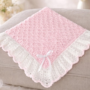 May include: A handmade, square baby blanket in soft pink with a white, scalloped lace border. A white satin ribbon is tied in a bow at the corner. The blanket is displayed on a neutral-colored cushion.