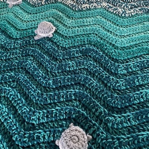 May include: A handmade crochet blanket in shades of teal, turquoise, and white, resembling ocean waves. The blanket features three-dimensional gray crochet sea turtles scattered across the surface, adding a whimsical touch to the aquatic theme.