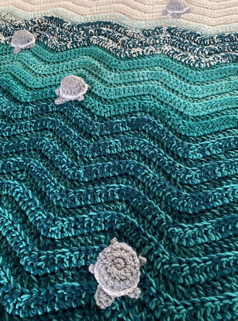 May include: A handmade crochet blanket in shades of teal, turquoise, and white, resembling ocean waves. The blanket features three-dimensional gray crochet sea turtles scattered across the surface, adding a whimsical touch to the aquatic theme.