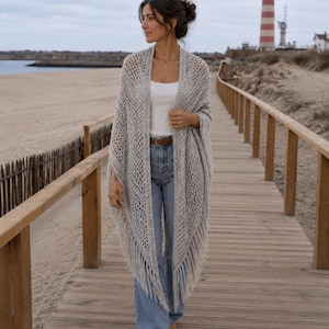 Pode incluir: Um xale cinza claro, de tecido aberto, com franjas é usado sobre uma blusa branca e jeans azuis. O xale é drapeado sobre os ombros. Um calçadão de madeira leva a uma praia com um farol ao fundo.