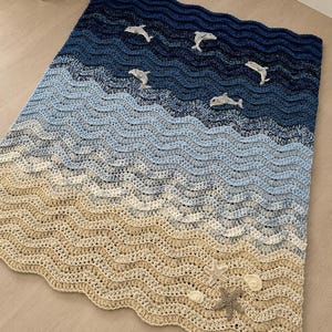 May include: A crocheted blanket depicting an ocean scene. The blanket features shades of blue and beige, with wave patterns and appliqued dolphins, starfish, and seashells. The blanket is designed to resemble a beach scene.