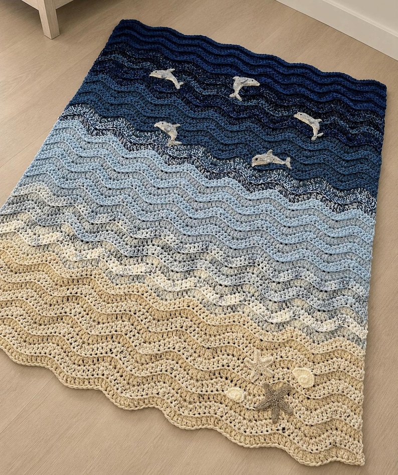 May include: A crocheted blanket depicting an ocean scene. The blanket features shades of blue and beige, with wave patterns and appliqued dolphins, starfish, and seashells. The blanket is designed to resemble a beach scene.