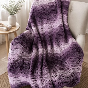 May include: A handmade, purple crochet blanket draped over a beige armchair. The blanket features a wave pattern with varying shades of purple and white. The blanket is made of soft yarn and is perfect for snuggling up on the couch.