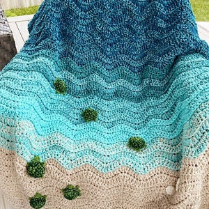 May include: A handmade crochet blanket with a beach theme. The blanket features a gradient of blues and beige, mimicking the ocean and sand. Small green crochet embellishments are scattered across the blanket, resembling sea turtles.