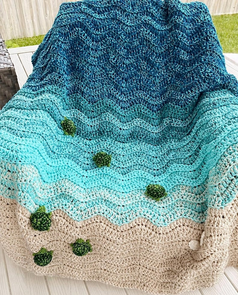 May include: A handmade crochet blanket with a beach theme. The blanket features a gradient of blues and beige, mimicking the ocean and sand. Small green crochet embellishments are scattered across the blanket, resembling sea turtles.