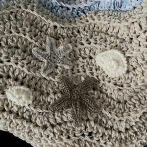 May include: Crocheted blanket in shades of beige, white, and blue, featuring a textured stitch pattern. The blanket is adorned with crocheted starfish and seashell appliqu&eacute;s, creating a coastal theme. The soft, handmade blanket is perfect for home decor.