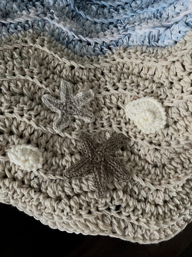 May include: Crocheted blanket in shades of beige, white, and blue, featuring a textured stitch pattern. The blanket is adorned with crocheted starfish and seashell appliqu&eacute;s, creating a coastal theme. The soft, handmade blanket is perfect for home decor.