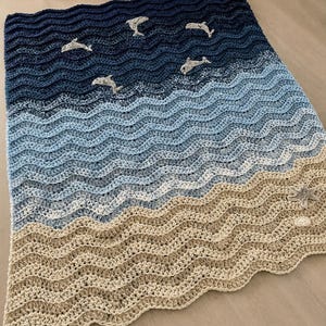 May include: A crocheted blanket with a wave pattern in shades of blue and tan, featuring several gray dolphin appliqu&eacute;s. The blanket's design evokes a beach scene, with the dolphins appearing to swim in the ocean.