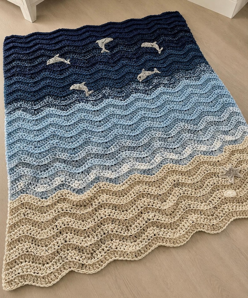 May include: A crocheted blanket with a wave pattern in shades of blue and tan, featuring several gray dolphin appliqu&eacute;s. The blanket's design evokes a beach scene, with the dolphins appearing to swim in the ocean.