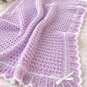May include: A soft, lavender crochet baby blanket with a scalloped white border and white ribbon accents. The blanket features a textured stitch pattern and is designed for comfort and warmth. A perfect handmade gift for a newborn.