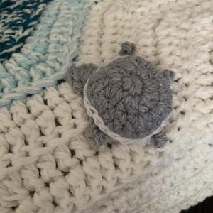 May include: A handmade crochet blanket in shades of white, teal, and light blue. A gray crocheted turtle with white trim is attached to the blanket. The blanket has a textured, woven appearance.