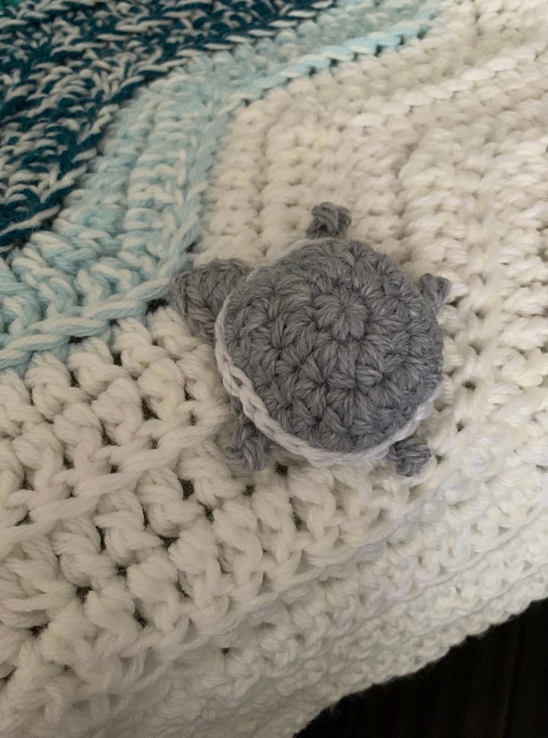 May include: A handmade crochet blanket in shades of white, teal, and light blue. A gray crocheted turtle with white trim is attached to the blanket. The blanket has a textured, woven appearance.
