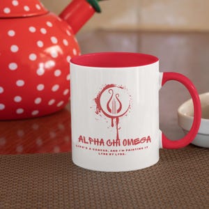May include: White ceramic mug with a red rim and handle. The mug features a red graphic of a lyre with a red circle and dripping paint. The text "ALPHA CHI OMEGA" is printed in red below the graphic. The text "LIFE'S A CANVAS, AND I'M PAINTING IT LYRE BY LYRE." is printed in red below the graphic.