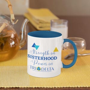 May include: A white ceramic coffee mug with a blue rim and handle. The mug features a blue, yellow and white triangle design and the text "Strength in Sisterhood Power in Tri Delta".