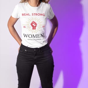 May include: White t-shirt with the text "REAL. STRONG. WOMEN. ALPHA CHI OMEGA" and a red fist with a gold sunburst around it. The numbers 18 and 85 are on either side of the fist.