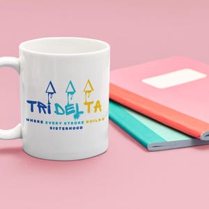 May include: White ceramic mug with a blue, yellow and white graphic of three arrows pointing upwards and the text "Tri Delta" and "Where every stroke builds sisterhood".